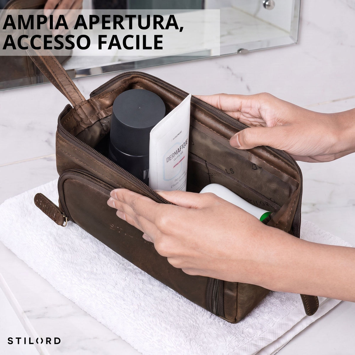 Necessaire in pelle marrone (zamora) aperto, interno con prodotti da bagno visibili, mani lo tengono su asciugamano